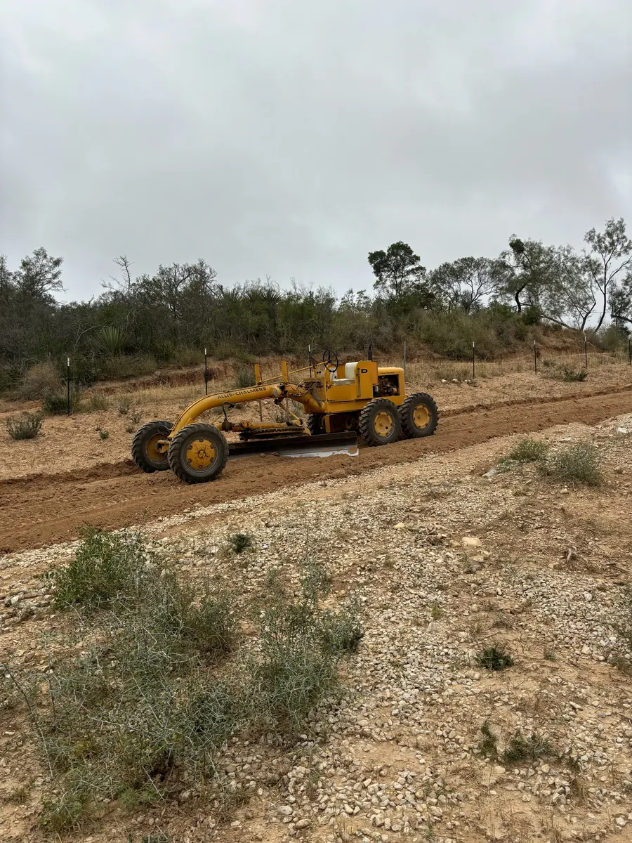Motor grader cutting the edge of a red dirt road through brush
