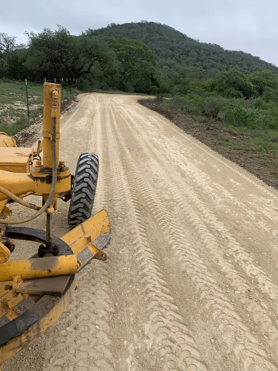 Motor grader blade beside a freshly graded road climbing toward a wooded hill