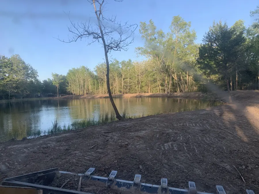 Pond holding water at the edge of woods, with freshly worked ground on the bank and a machine bucket edge in front