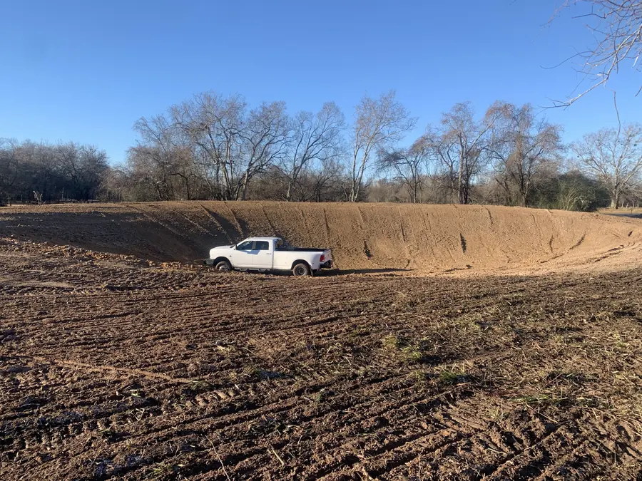 Freshly dug earthen tank with a white pickup parked inside, bare trees behind